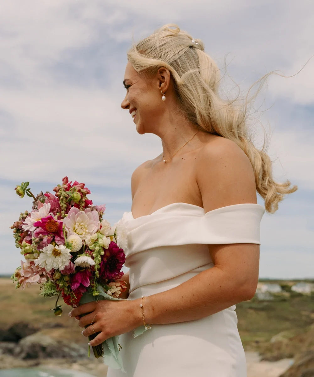 bride holdinBride wearing delicate gold pearl jewellery with an off-the-shoulder wedding dress and colourful bouquet on the Cornish coast.g white rose bouquet wearing delicate pearl bracelet, black and white wedding photography