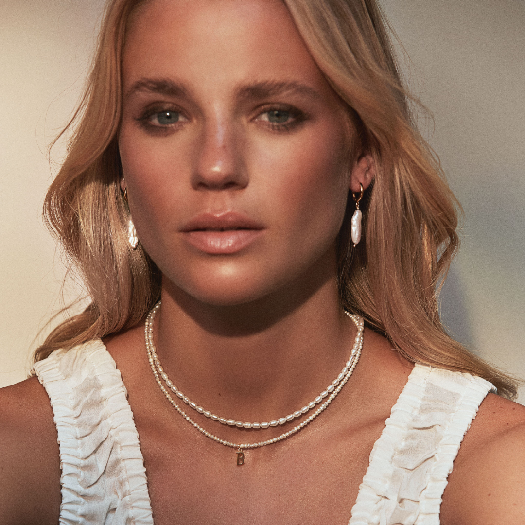 Gold seed pearl choker layered with a gold small pearl initial letter choker around her neck around a neck of a blonde woman wearing gold baroque pearl shard hoop earrings in her ears