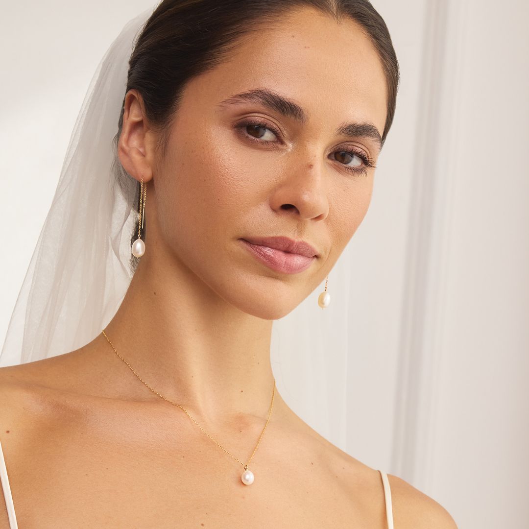 Woman wearing a gold pearl pendant necklace with matching pearl threader earrings against a white background.