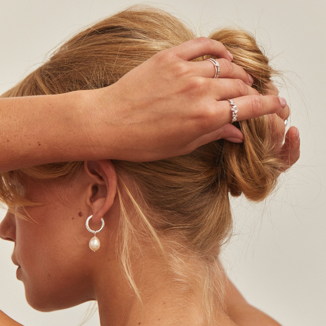 A blonde woman wearing a silver diamond style large pearl drop hoop earring in her ear lobe holding her hair back, with a silver diamond style baguette ring on her finger