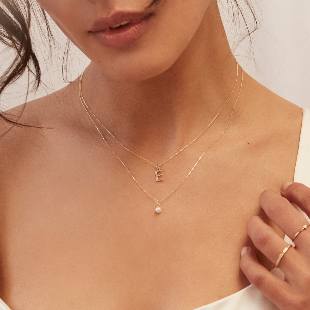 Close up of a woman wearing a gold single pearl necklace and a gold E letter necklace