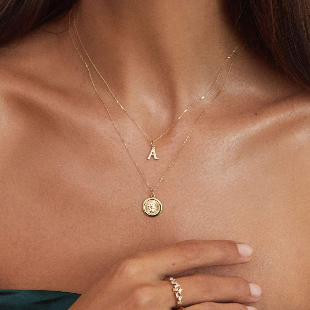Close up of a solid gold curve initial letter necklace 'A' paired with a gold St Christopher medallion necklace around the neck of a brunette woman wearing a gold dimond style baguette ring on her finger 