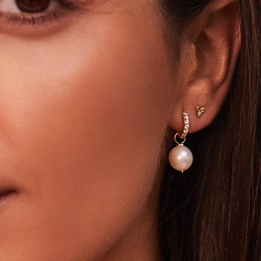 A gold diamond style pyramid stud earring and a gold diamond style large pearl drop hoop earring in an ear lobe of a brunette woman