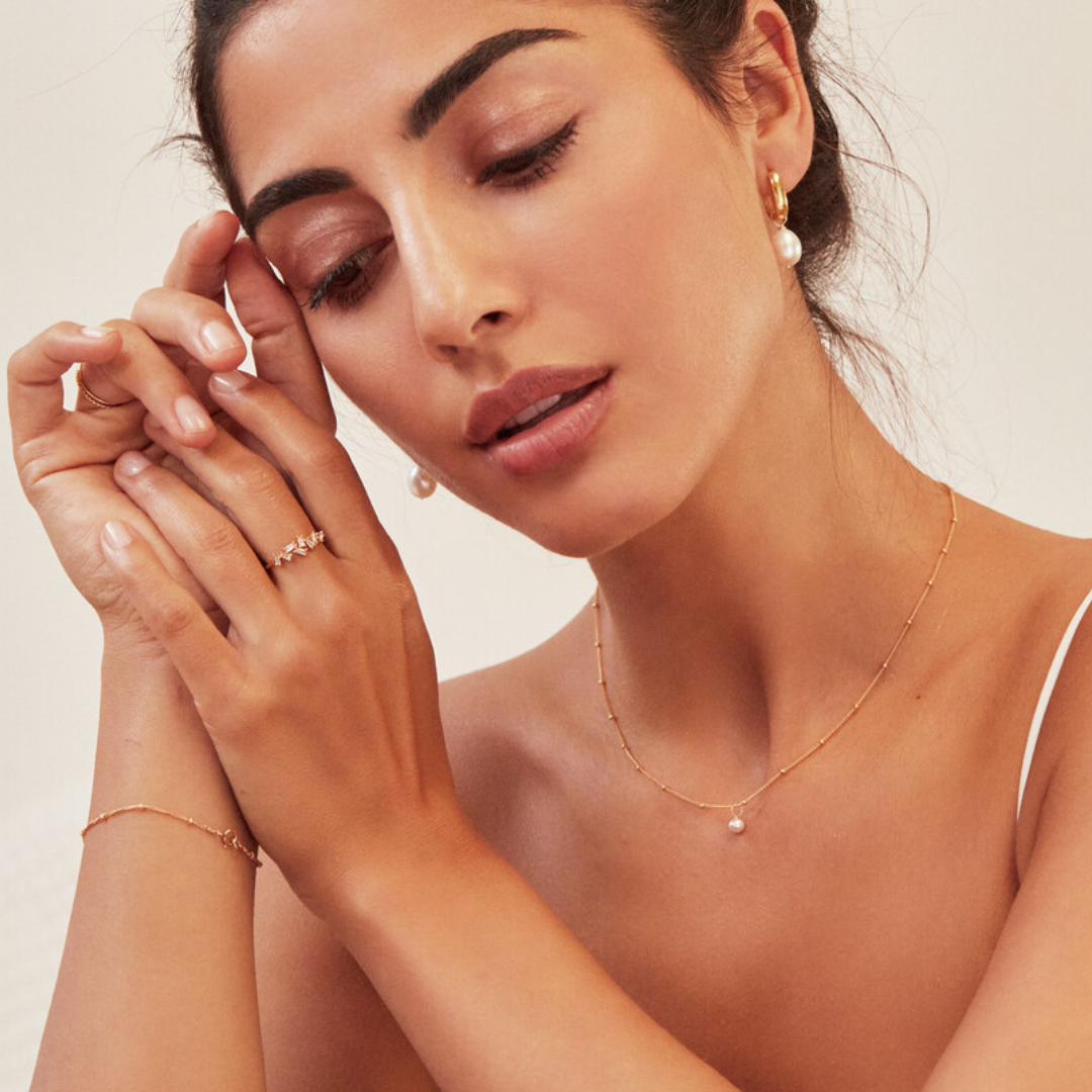Woman wearing a gold single pearl satellite necklace around her neck, gold thick squared hoop pearl drop earrings in her ears and a gold diamond style baguette ring on her finger clasping her hands together next to her face