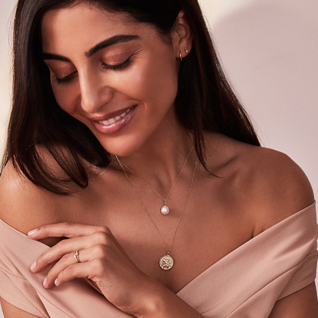A smiling woman looking down wearing a gold wave huggie hoop earring and a stud in one ear lobe and a gold large single pearl necklace around her neck