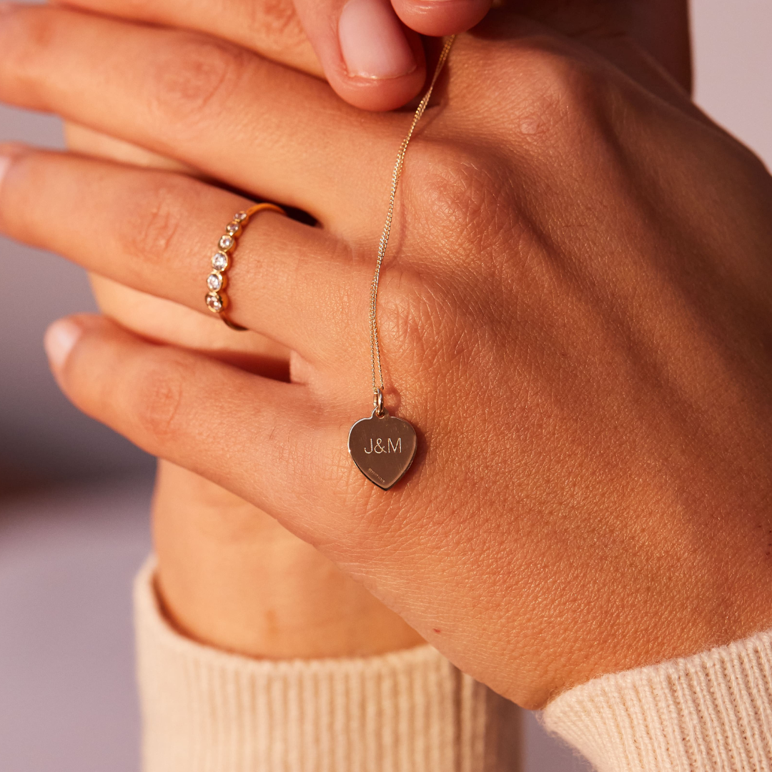 Other side of gold small heart st christopher necklace with the initials 'J&M' held by a woman over her knuckles, also wearing a gold diamond style round eternity ring on her finger