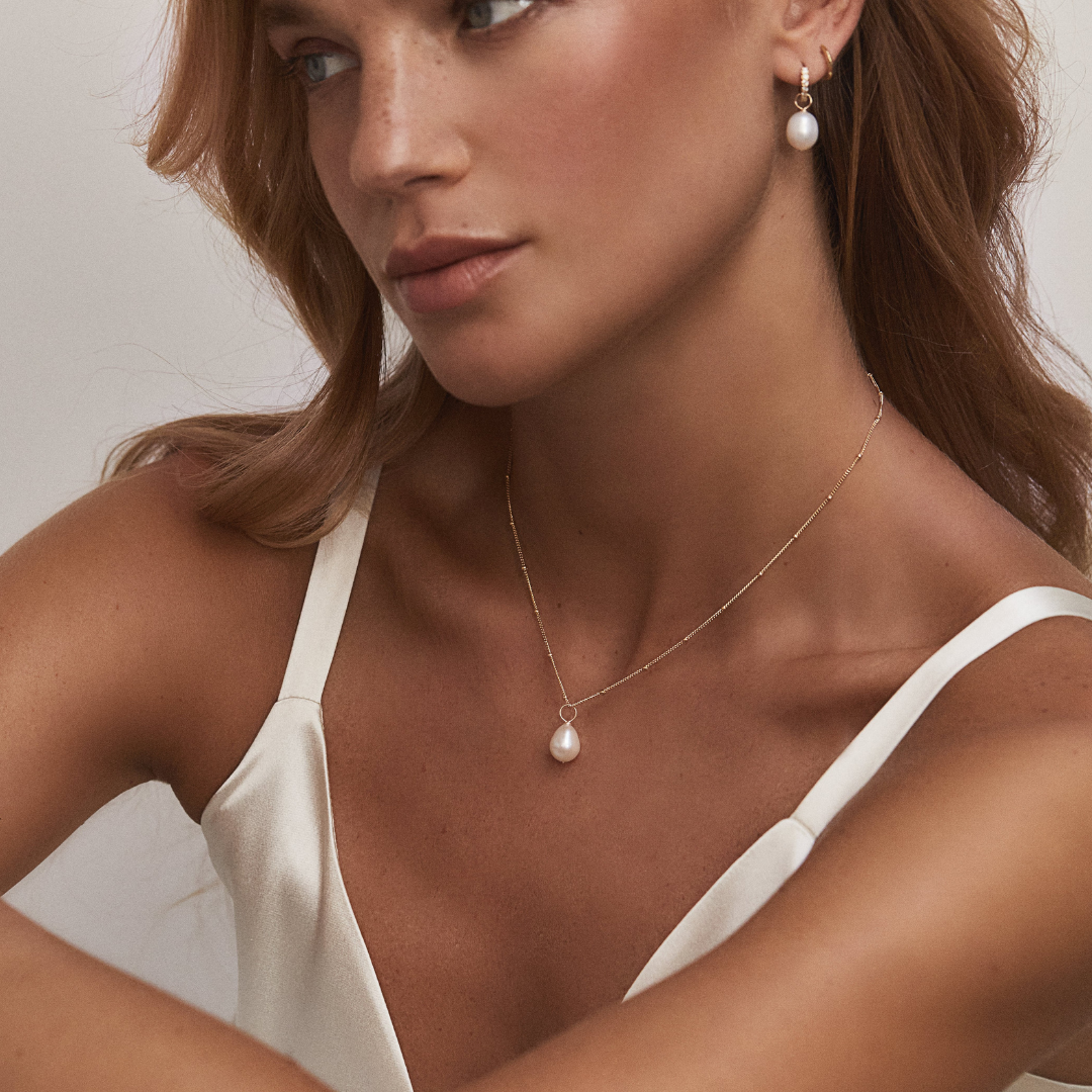 Gold diamond style large pearl drop hoop earring in one ear lobe of a blonde woman with the gold large pearl satellite necklace around her neck 