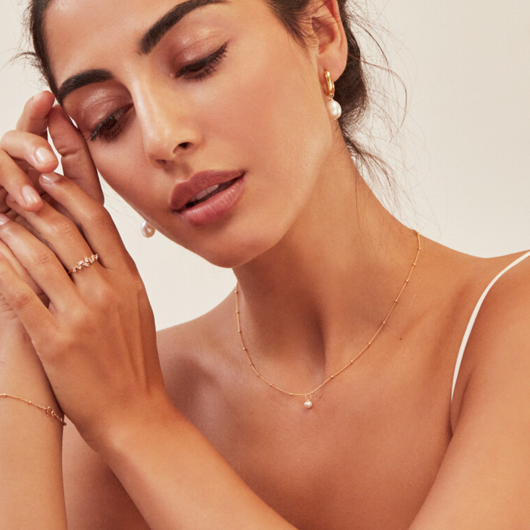 Brunette woman wearing a gold diamond style baguette ring on her finger and a gold satellite single pearl choker around her neck with her head tilted to the side looking down