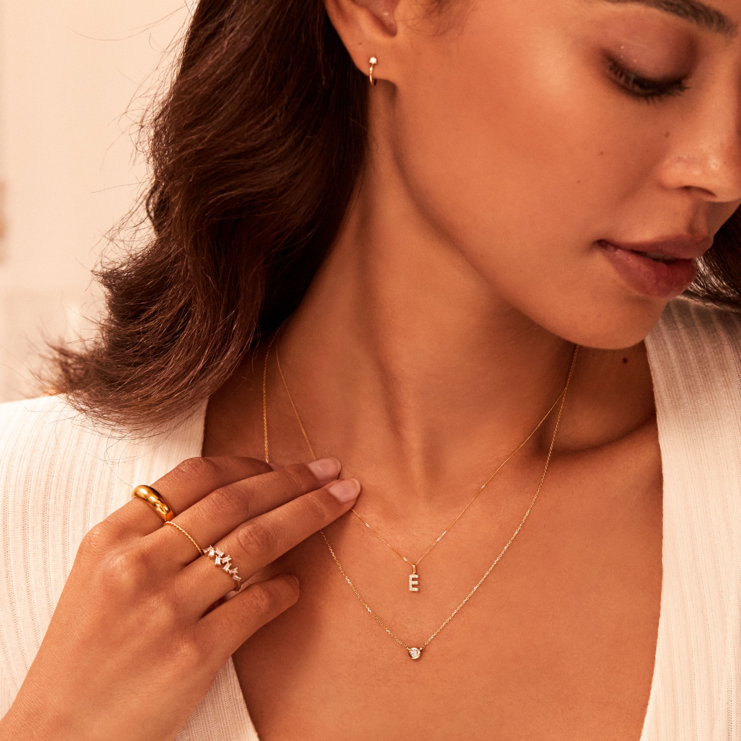 A woman wearing a gold diamond style lobe hoop stud earring in her ear lobe, a gold single diamond style necklace layered with a gold chain necklace with a solid gold individual initial charm 'E' around her neck