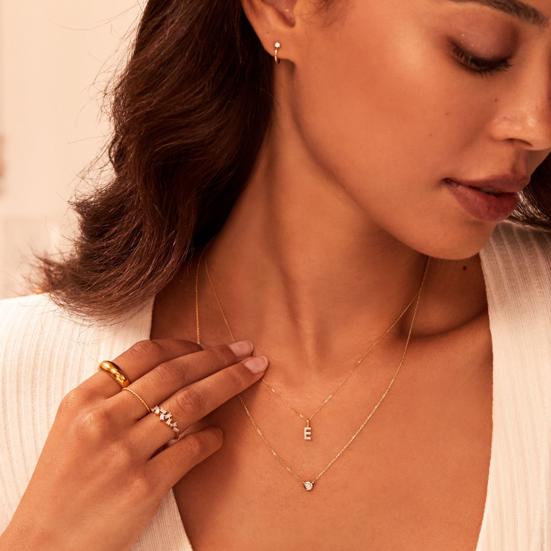 a woman wearing a gold thin twisted stacking ring on one finger, a gold diamond style baguette ring on another finger and a gold plain dome ring on another finger with a single diamond choker and 'E' letter necklace around her neck