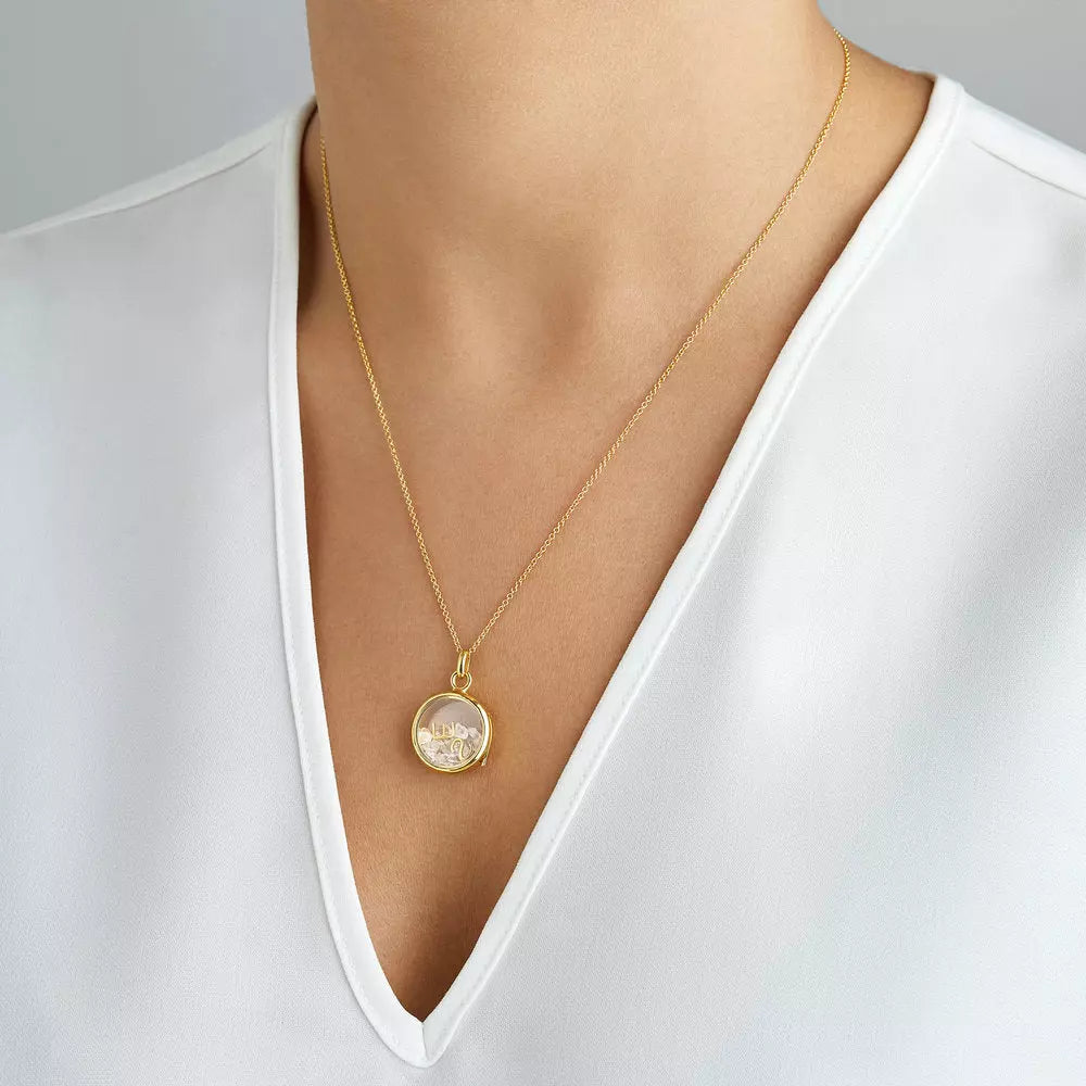Gold glass gemstone locket with transparent white gemstones and some gold letters around a neck with a white V neck tshirt