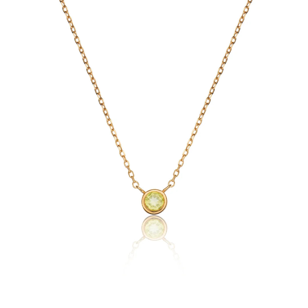 A delicate gold chain necklace with a small round bezel-set yellow-green gemstone pendant, displayed on a clean white background with a soft reflection below.