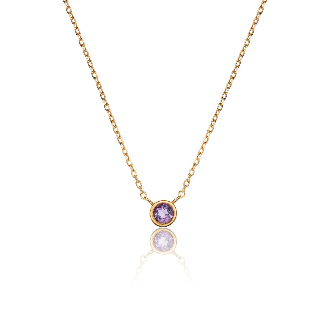 A delicate gold chain necklace featuring a single bezel-set round purple gemstone pendant, displayed against a clean white background with a soft reflection below.