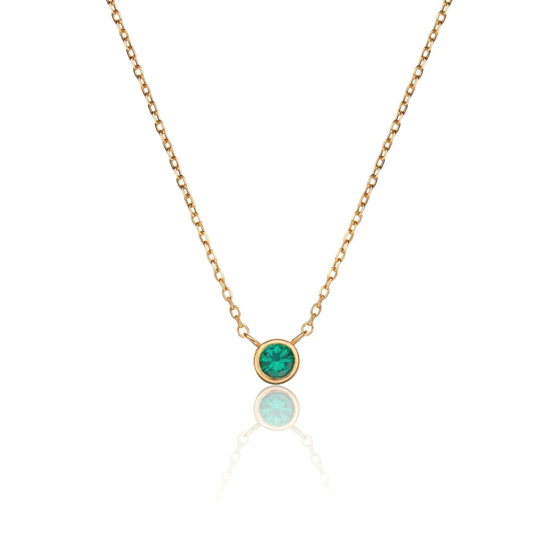 A delicate gold chain necklace with a small round bezel-set green stone pendant, displayed against a clean white background with a soft reflection below.