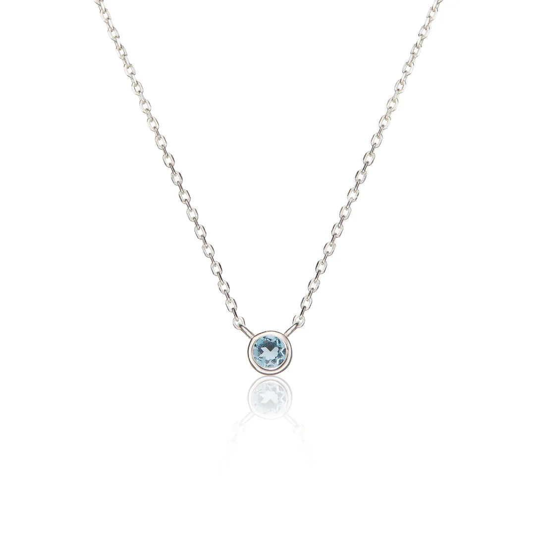 A delicate silver chain necklace featuring a small bezel-set round light blue gemstone pendant, displayed on a clean white background with a soft reflection below.