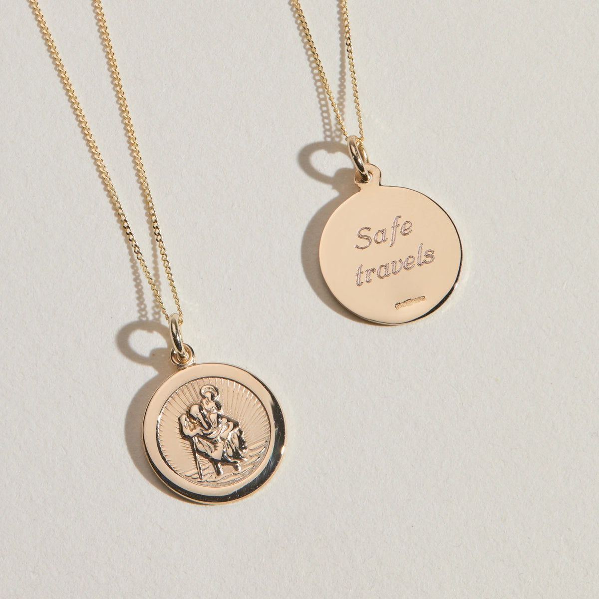 Two gold round pendant necklaces on a light beige background. One pendant is engraved with 'Safe travels' and the other features an embossed figure of St Christopher, both on delicate chains.