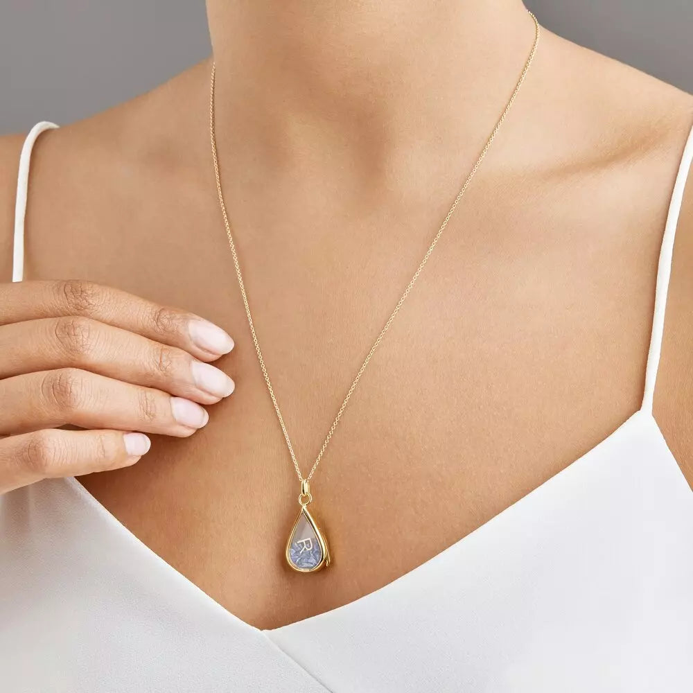 Gold glass gemstone teardrop locket with blue gemstones and a gold R around neck with a white top