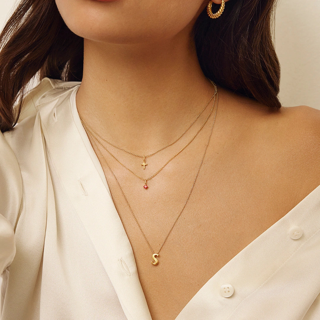 Close-up of a woman wearing layered gold necklaces, including a delicate chain necklace with an initial 'S' pendant, a small cross pendant, and a fine chain with a tiny red gemstone pendant, styled with a white satin blouse.