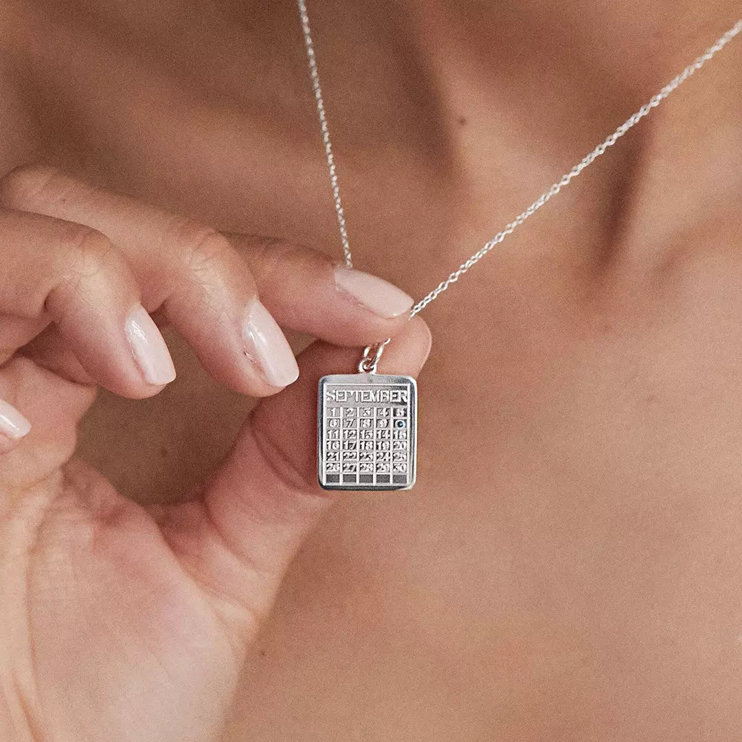 Close up of silver special date calendar necklace 'September' with a gemstone on number '10' around the neck of a woman holding it up with her hand