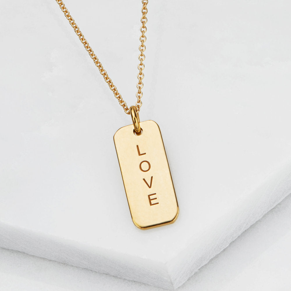Gold personalised tag necklace with 'LOVE' engraved on it on a marble surface