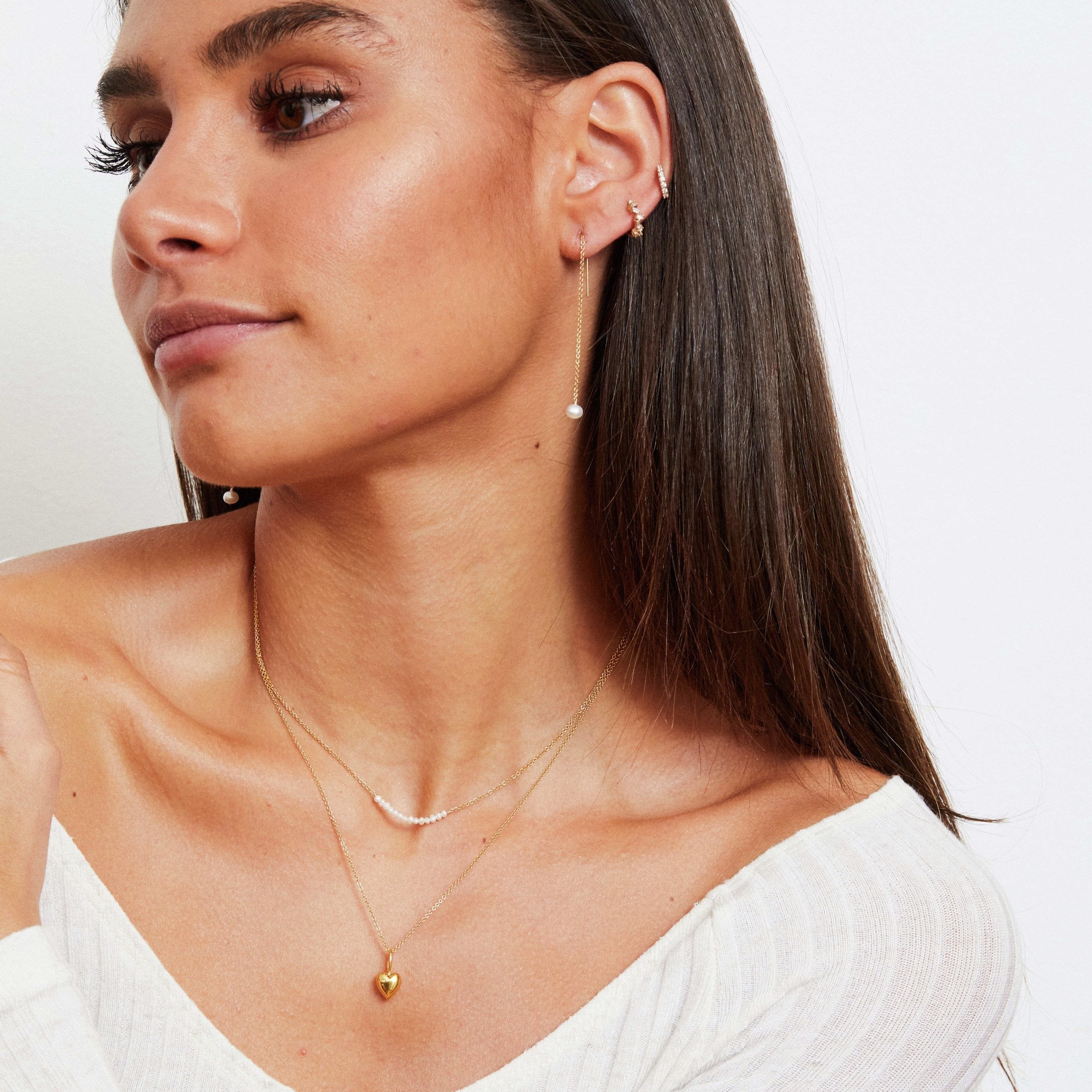 A woman wearing two gold diamond style baguette huggie hoop earrings with a pear thread earring, a gold pearl cluster necklace and a gold heart pendant necklace around her neck