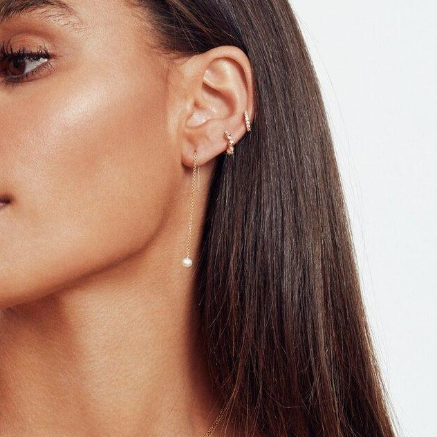 A gold pearl drop ear threader and two gold diamond style baguette huggie hoop earrings in one ear lobe of a brunette woman