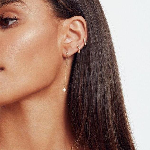 A gold pearl drop ear threader and two gold diamond style baguette huggie hoop earrings in one ear lobe of a brunette woman