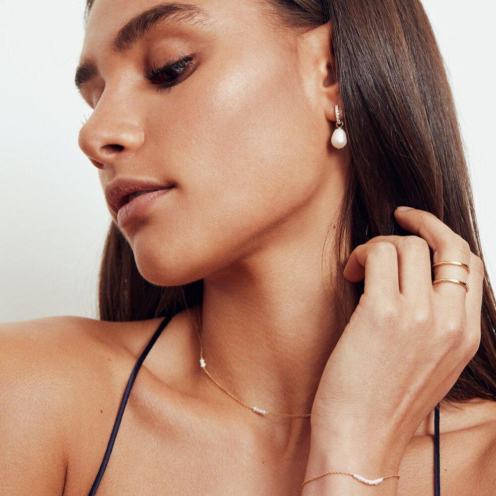 A brunette woman looking to the side wearing a gold diamond style large pearl drop earring in her ear lobe, a gold mini pearl choker around her neck, a gold small pearl cluster bracelet around her wrist and a gold double band ring on her middle finger