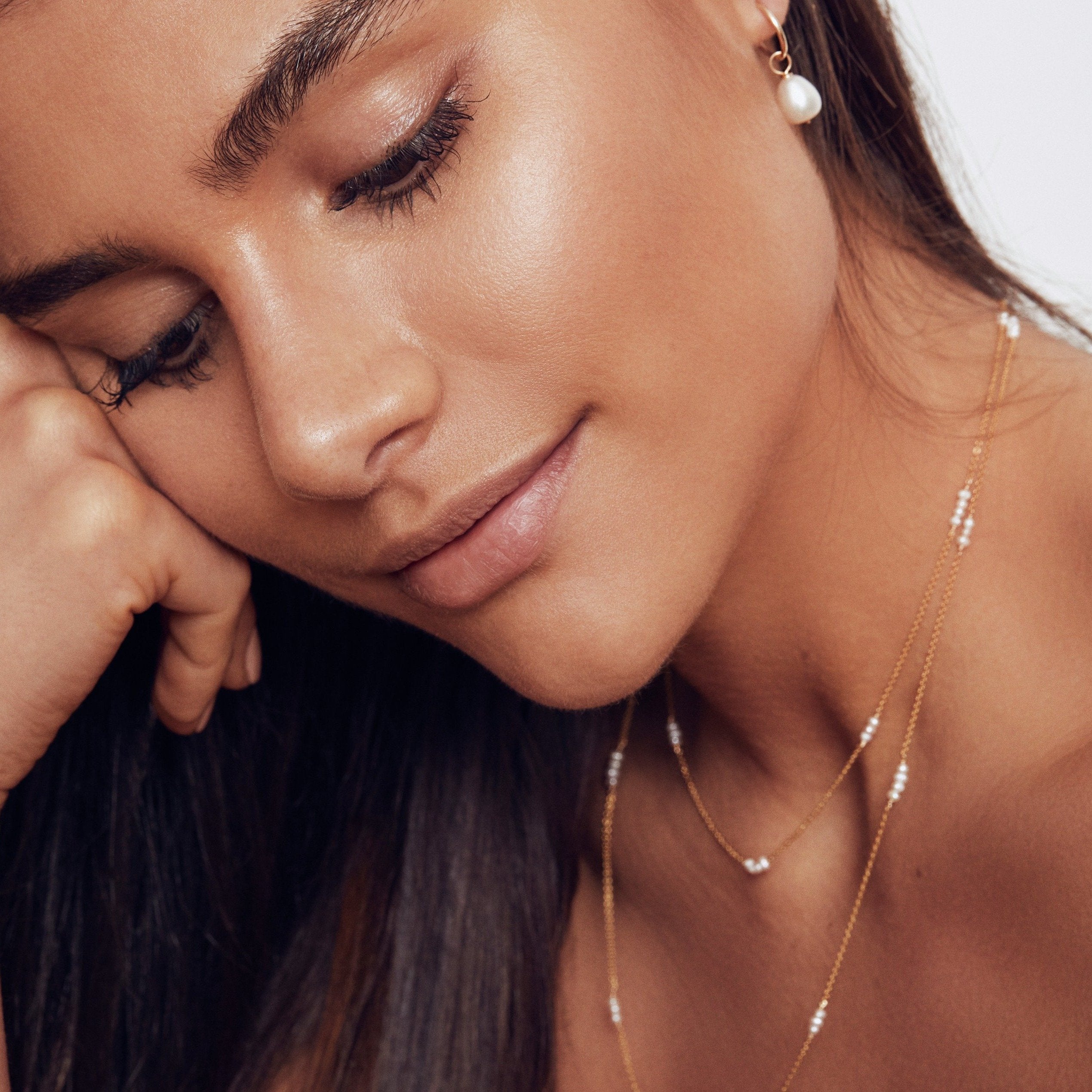 Gold layered mini pearl necklace around the neck of a brunette woman wearing a pearl earring in one ear lobe, her head resting on her knuckles