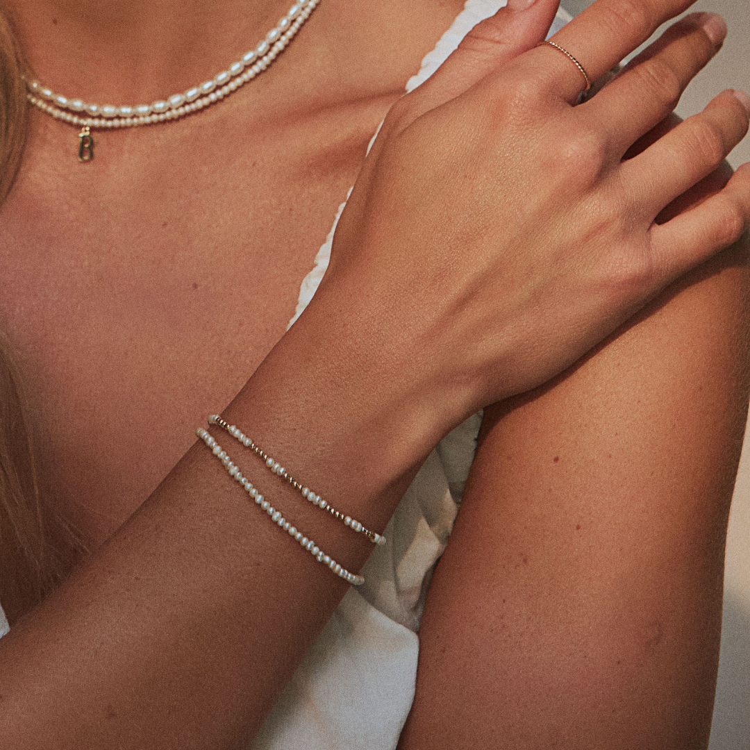 Close up of woman wearing a gold beaded mini pearl bracelet on her wrist layered with a gold small pearl bracelet and wearing a Bl letter necklace and gold seed pearl choker around her neck