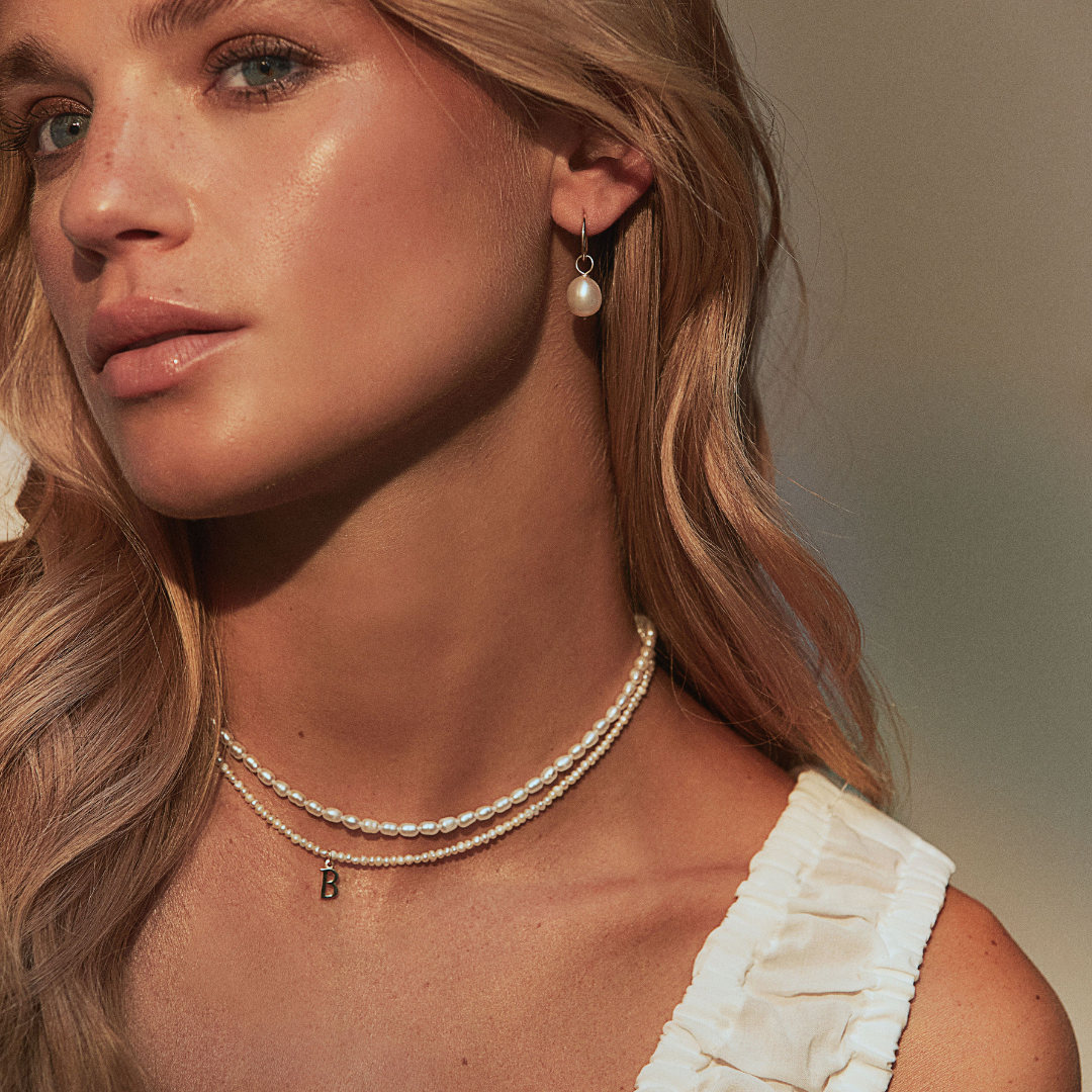 Gold small pearl initial letter choker around the neck of a blonde woman looking to the side also wearing a gold seed pearl choker around her neck and a pearl earring in her ear lobe