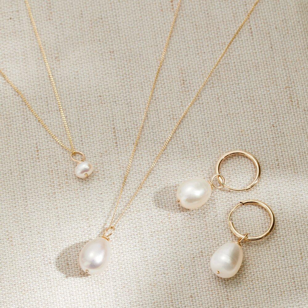 Gold single pearl necklace, gold large single pearl necklace and gold plain huggie pearl drop hoop earrings on a woven surface  