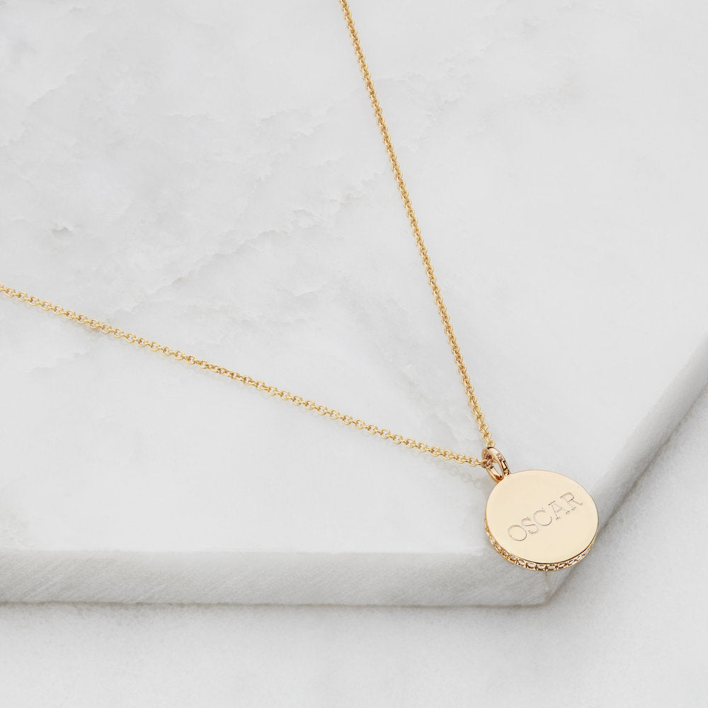 Gold small diamond style disc necklace with 'OSCAR' engraved on it on a marble surface