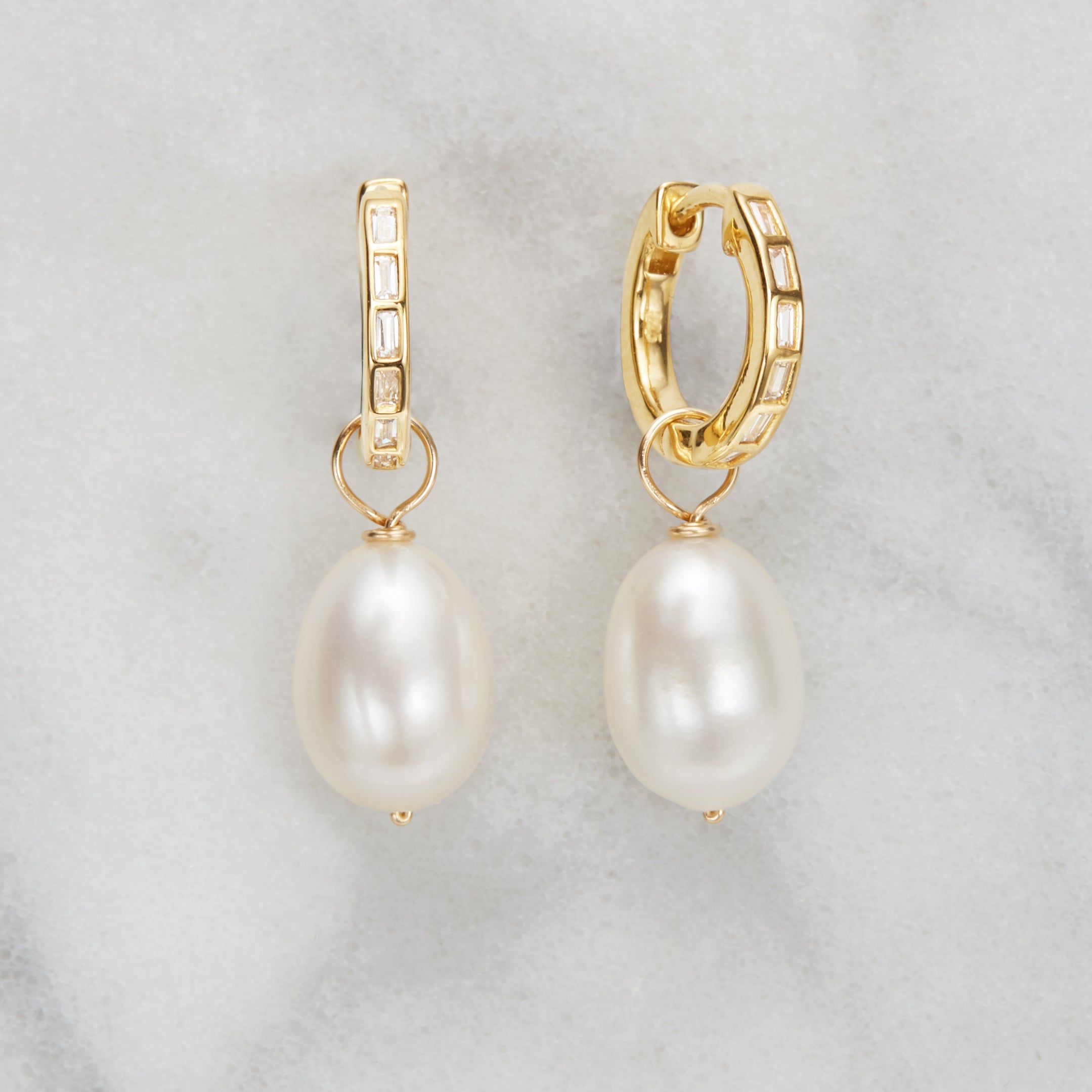 A pair of gold diamond style baguette pearl drop hoop earrings on a marble surface