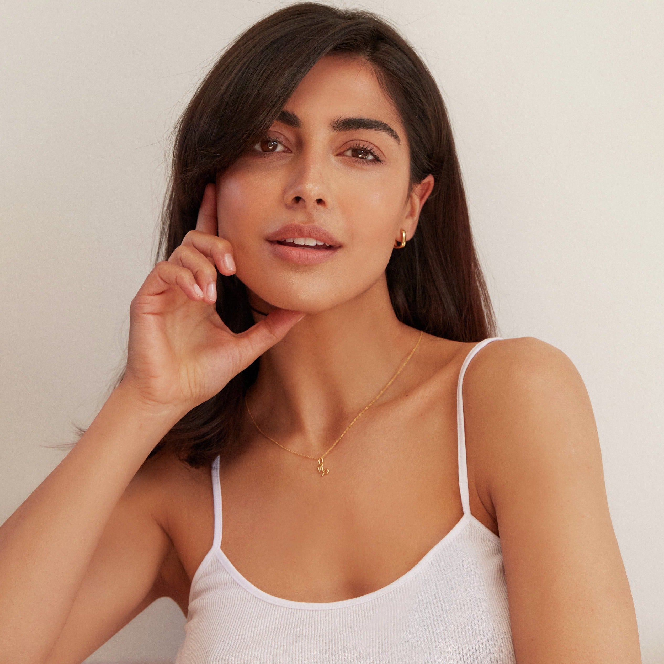 A gold thick squared hoop earring in an ear lobe of a brunette woman wearing a white strappy top with her chin resting on her thumb