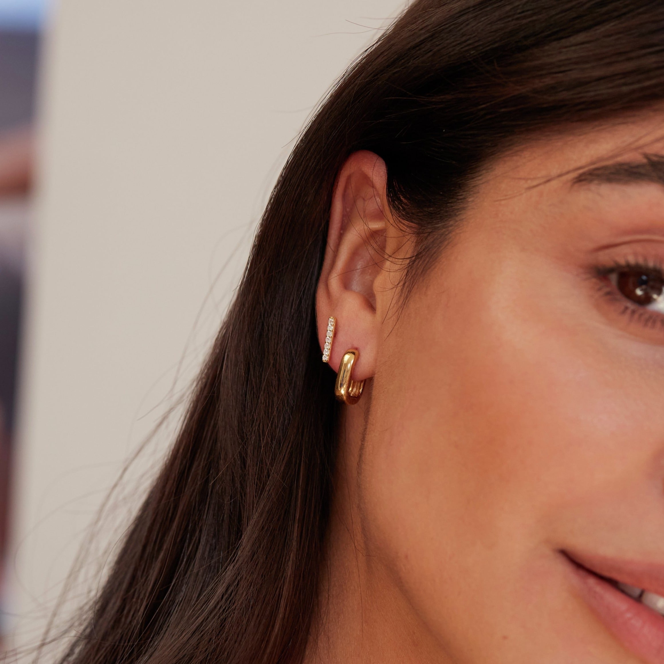 Close up of a gold diamond style bar stud earring and gold thick squared hoop earringin one ear lobe of a brunette woman