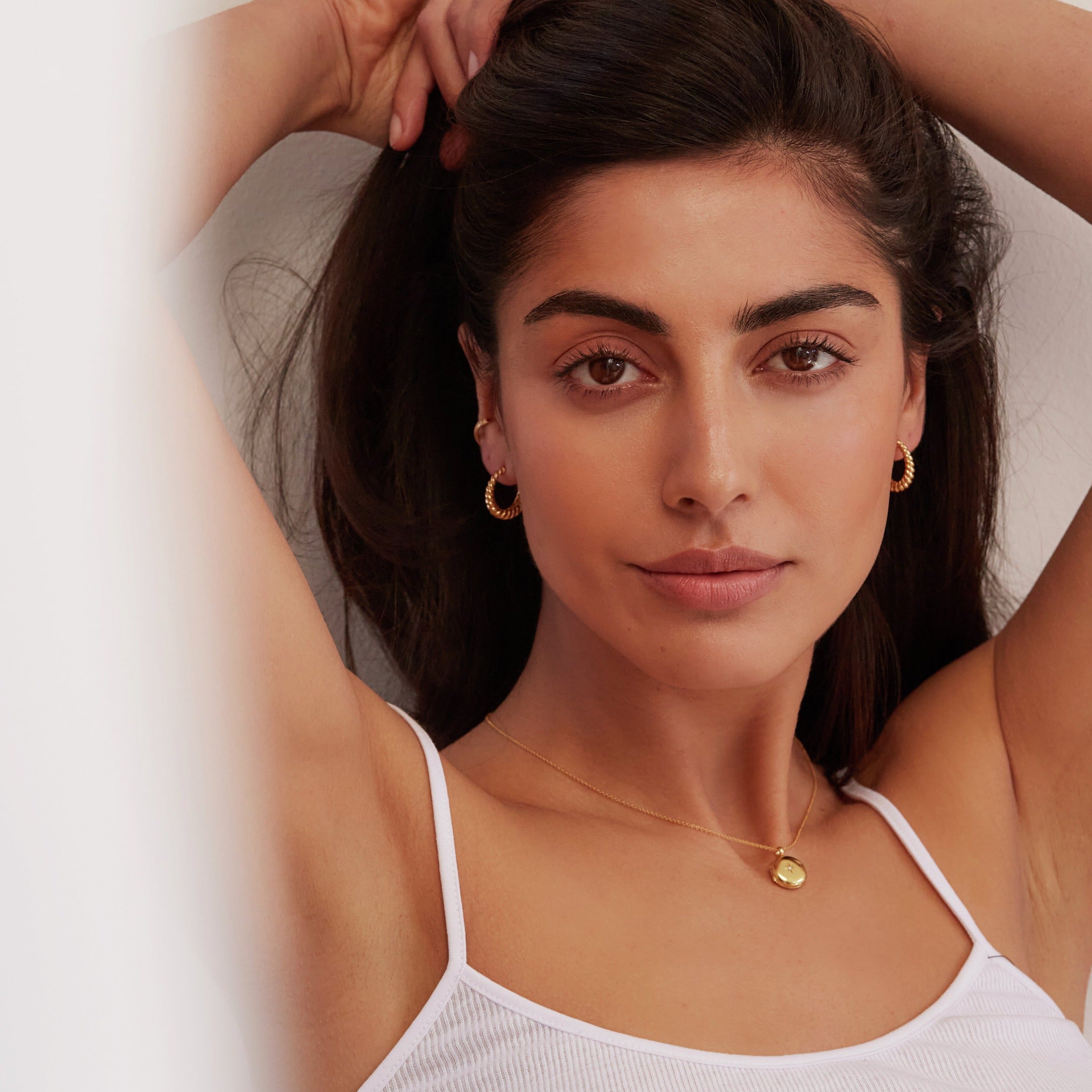 Gold large twisted rope hoop earrings worn by a brunette woman holding her hair back with her hands also wearing a gold small round diamond locket necklace