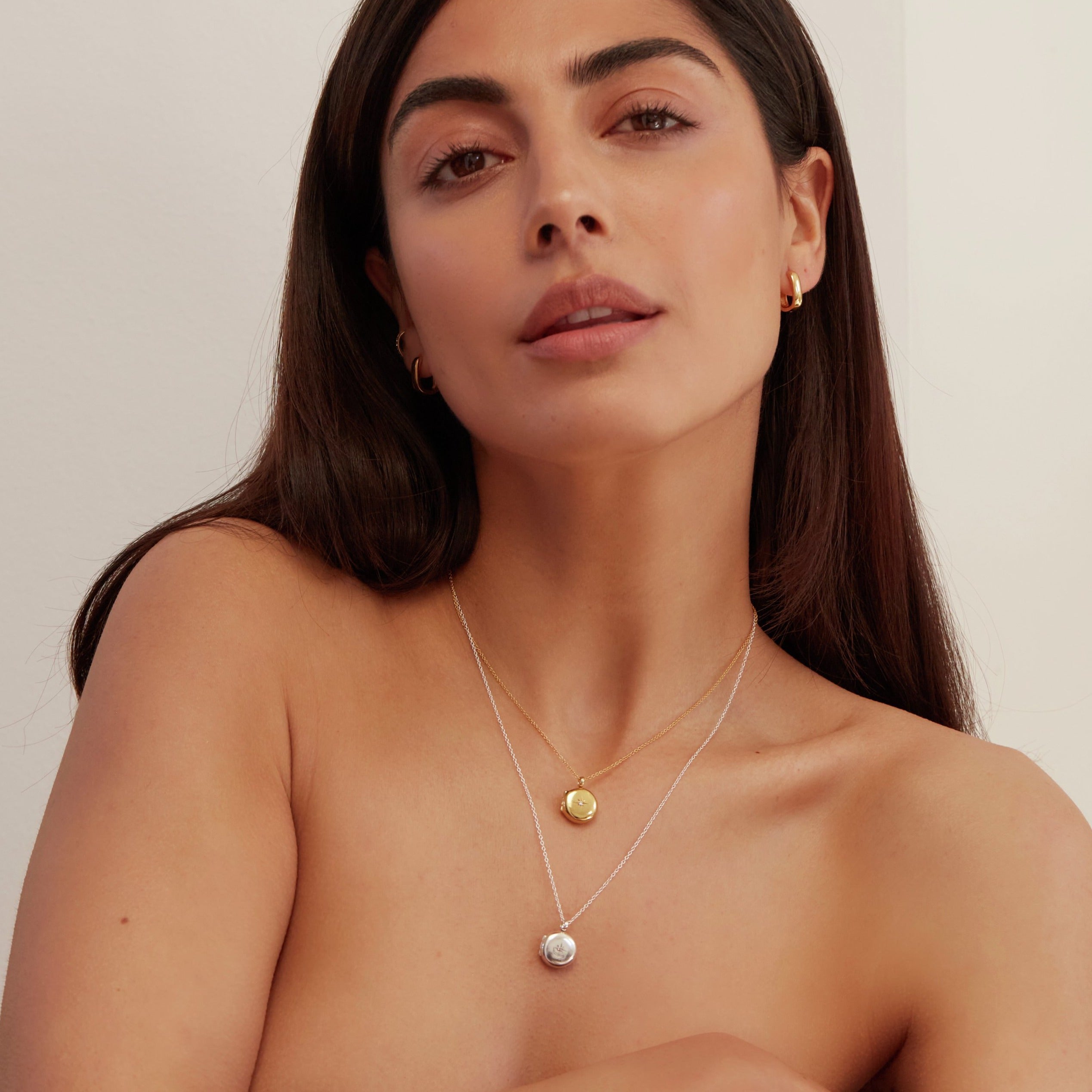 Woman wearing a silver small round diamond locket necklace layered with a gold small round diamond locket necklace around her neck and gold square edge hoop earrings in her ear lobes
