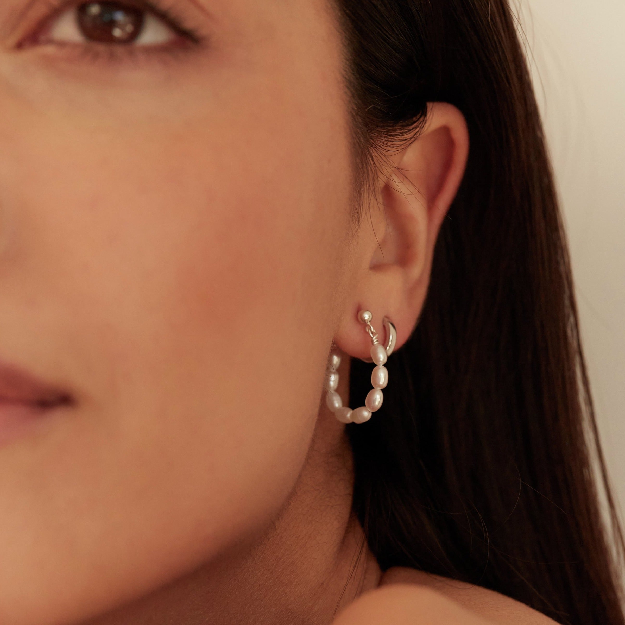 Silver seed pearl hoop earring and silver huggie hoop earring in an ear lobe of a dark haired woman