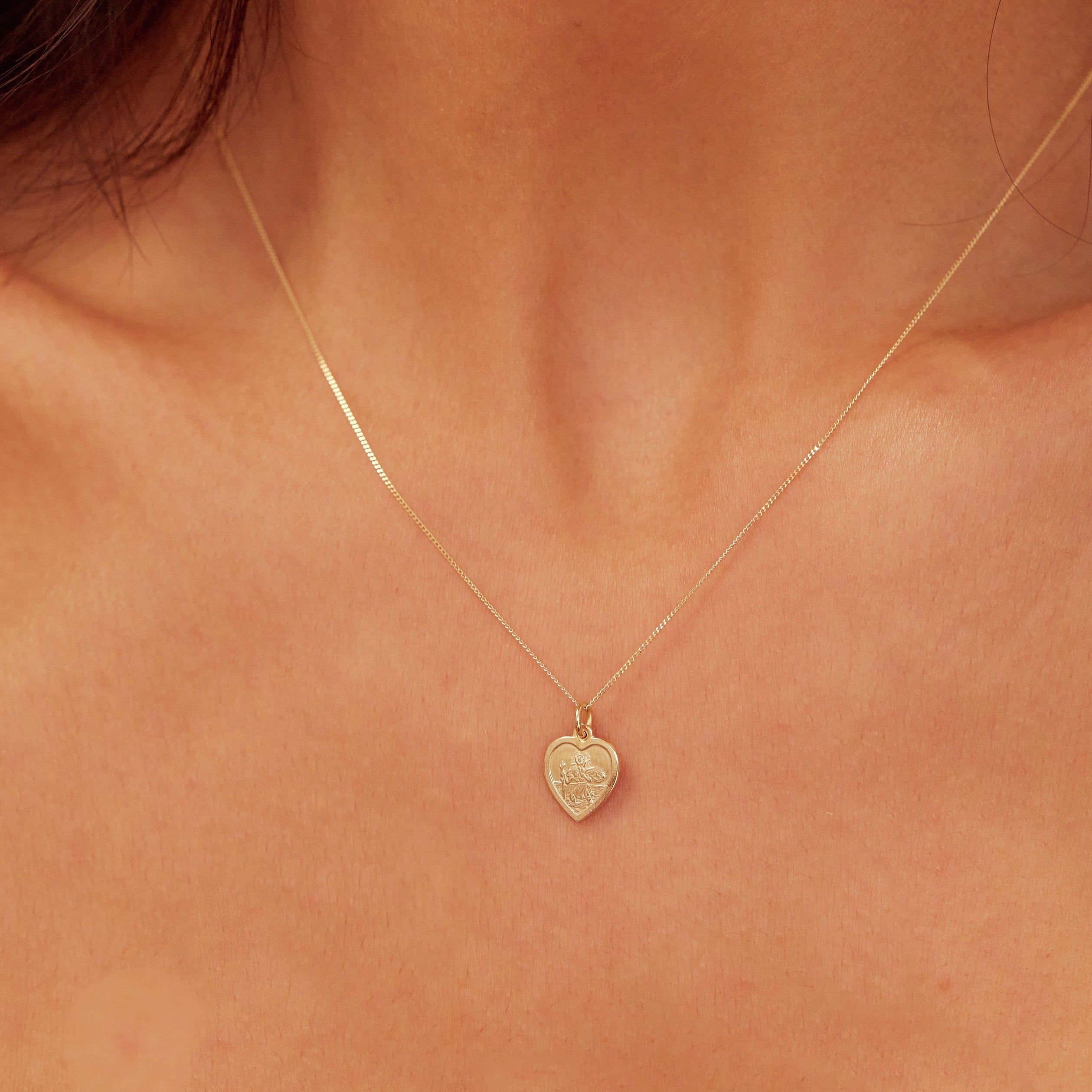 Close up of gold small heart st christopher necklace around the neck of a brunette woman