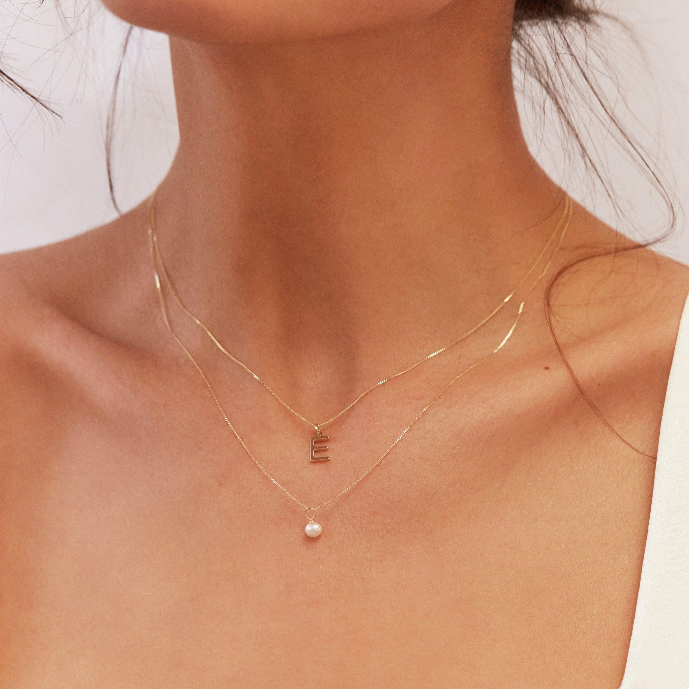 Gold single pearl necklace layered with a gold E letter necklace around a neck close up