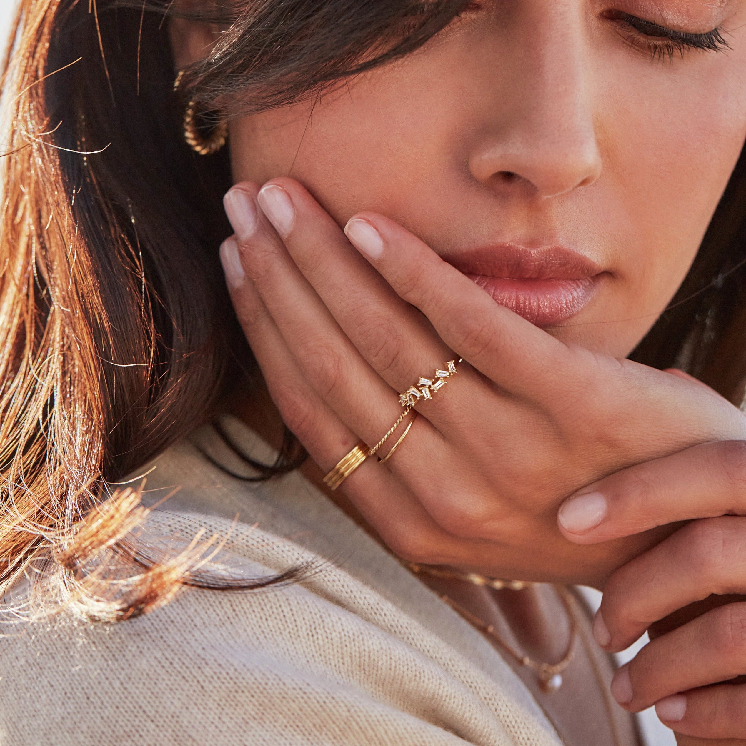A brunette woman wearing a gold thin twisted stacking ring, a gold diamond style baguette ring, a gold thin plain stacking ring and a gold thick triple band ring on her fingers
