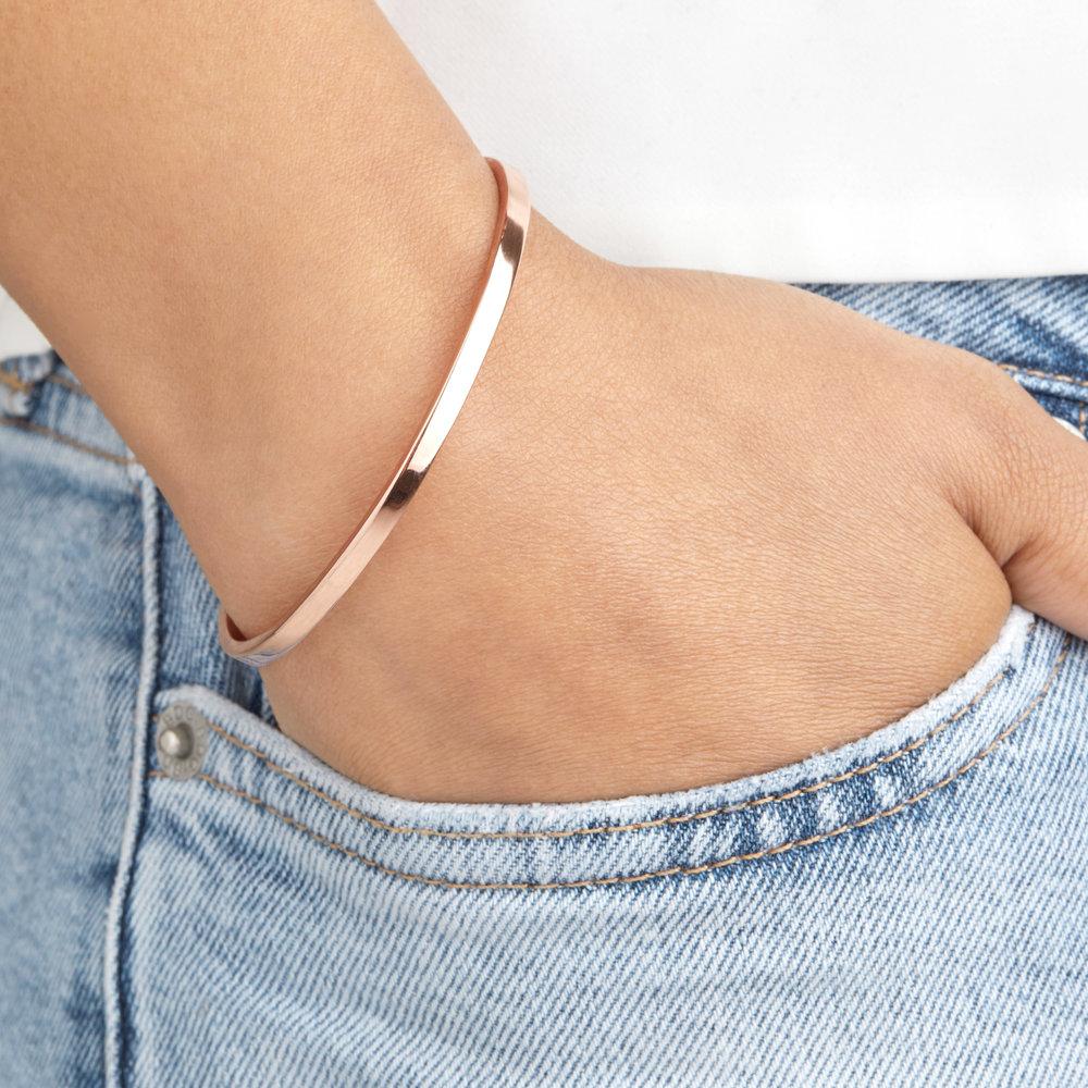 A rose gold thin engraved bangle worn on a wrist with the hand in a blue denim pocket