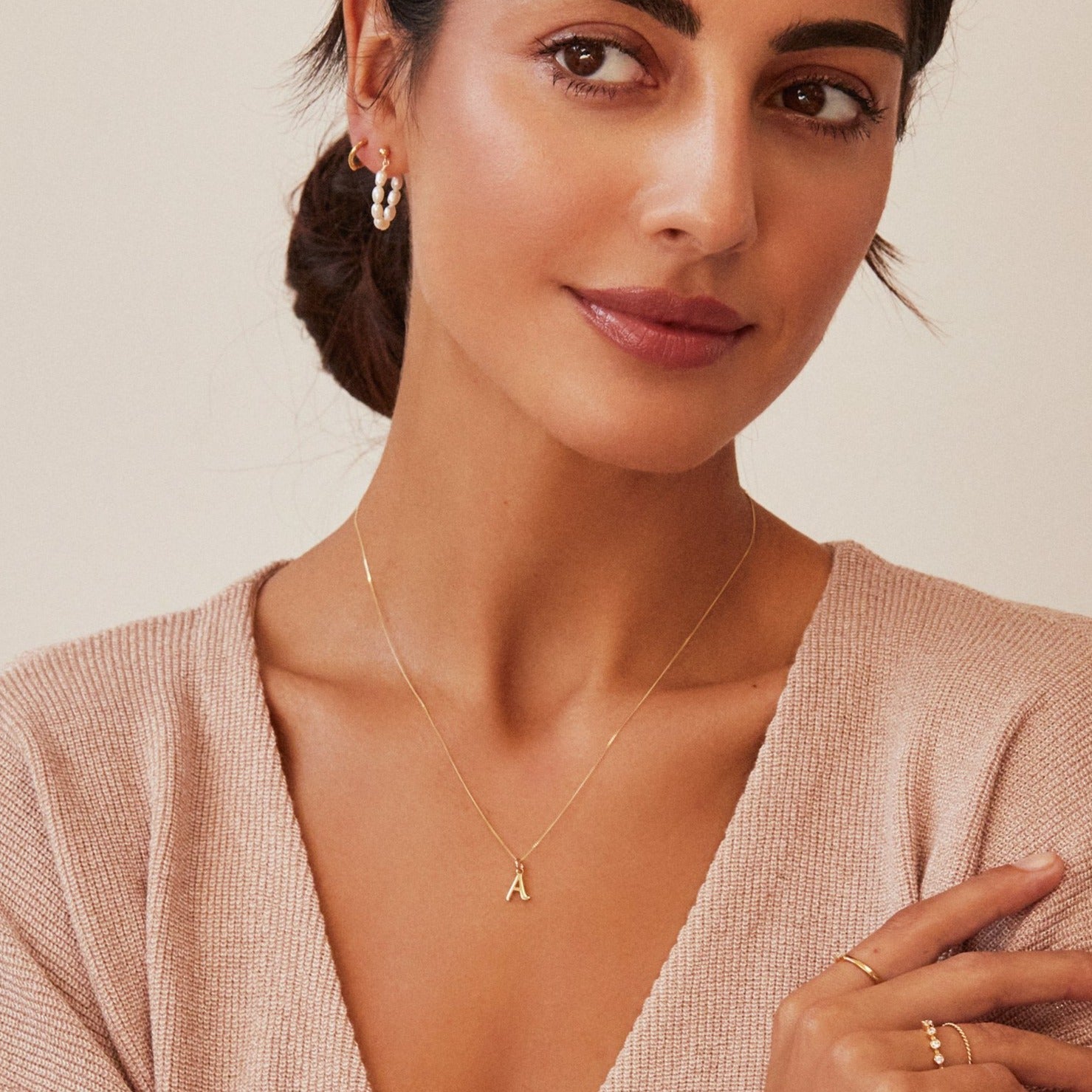 A woman wearing a gold curve initial letter necklace around her neck with a brown knitted jumper and gold seed pearl hoop earring in one ear