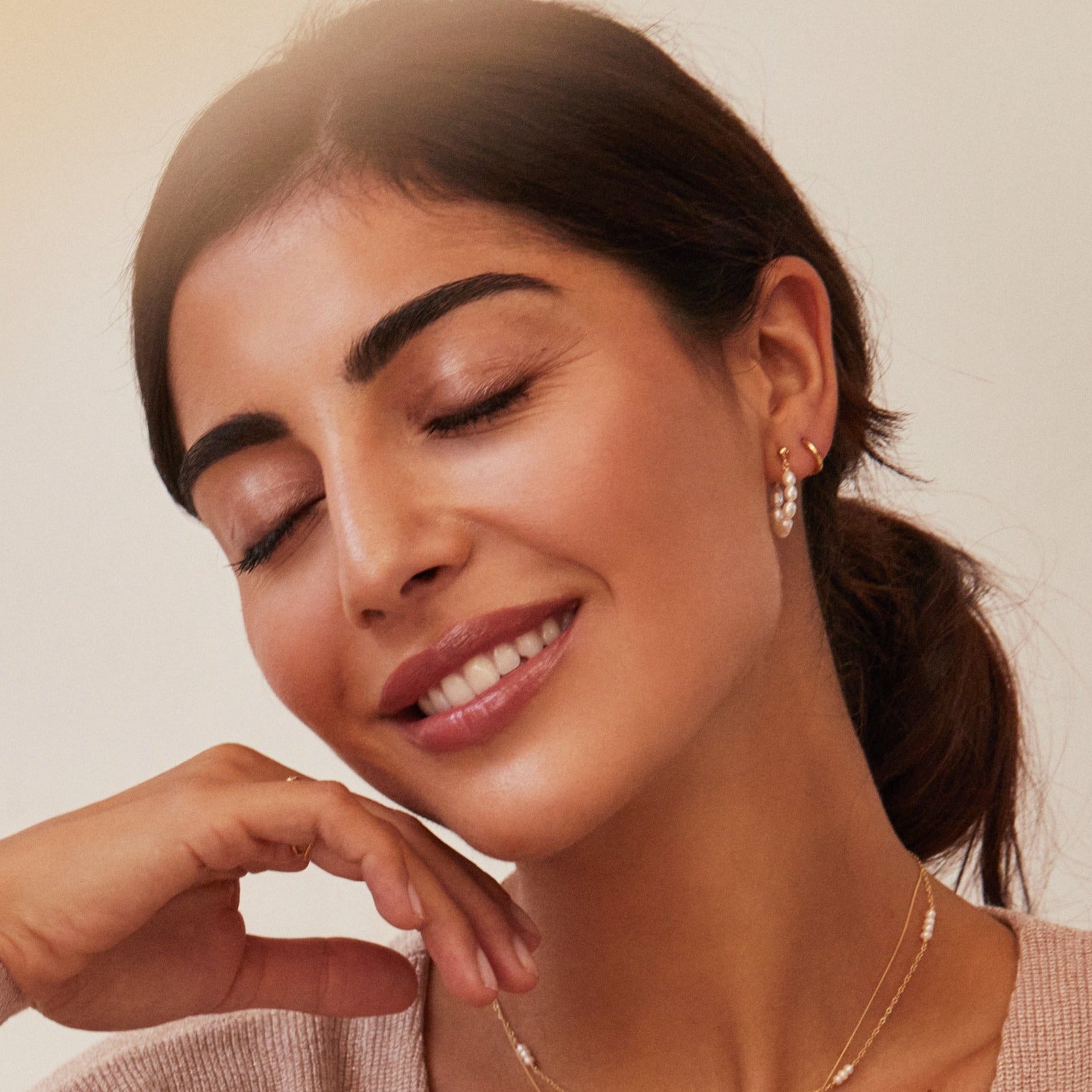 A woman wearing a gold seed pearl hoop earring and gold huggie hoop earring in one ear lobe and a pearl necklace around her neck with her eyes closed and smiling