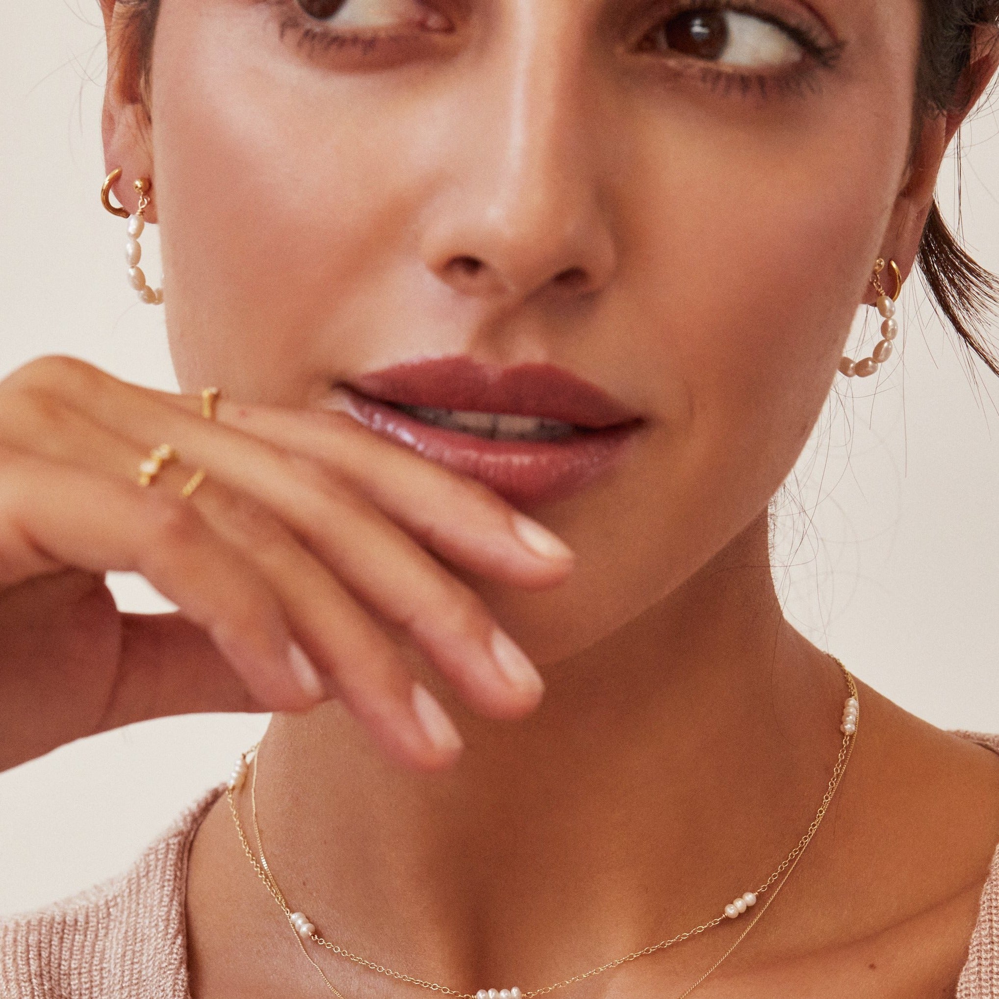 Gold seed pearl hoop earring and a gold hoop earring in a brunette woman's ear lobe, with her fingers resting on her lips, also wearing a gold mini pearl choker around her neck