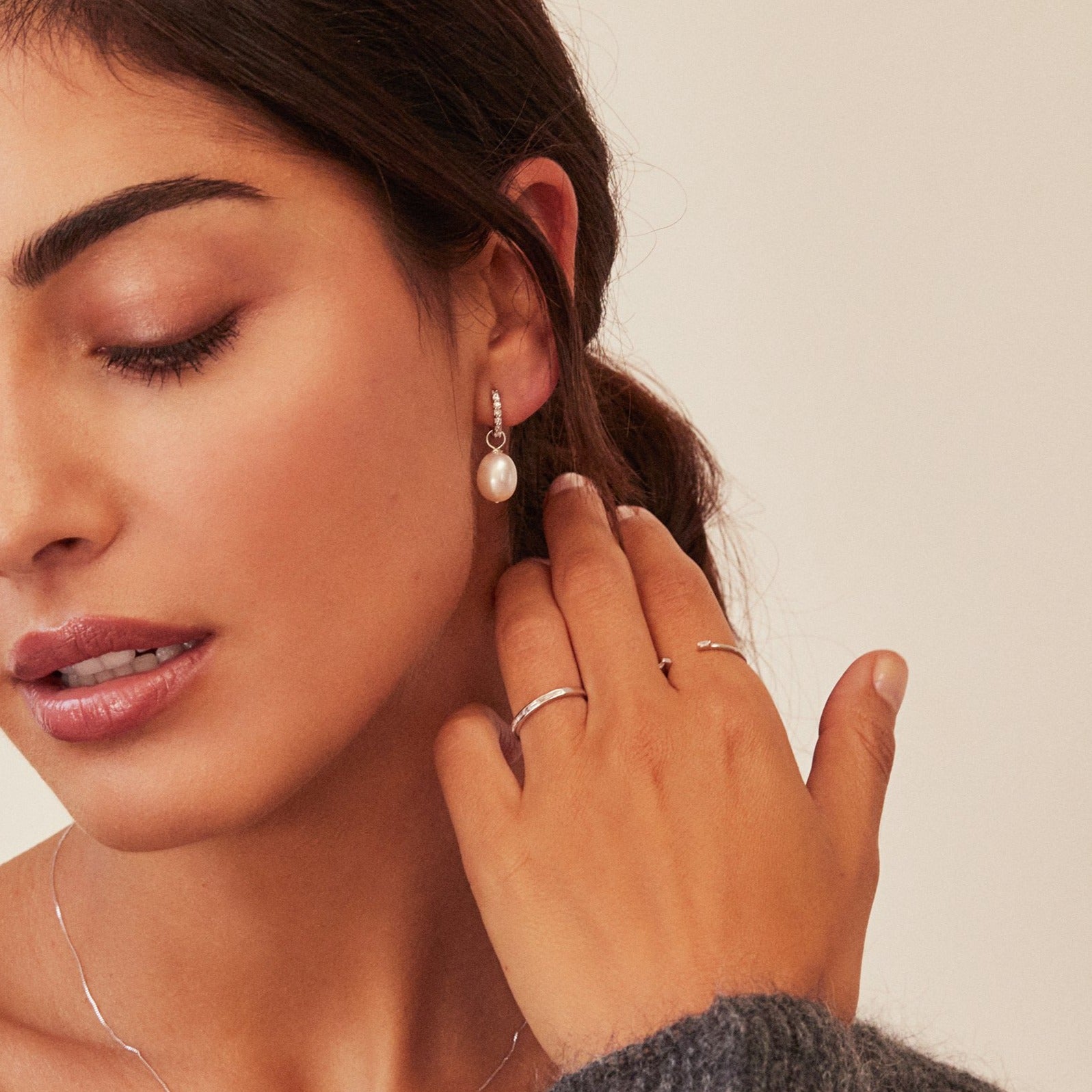 A brunnette woman wearing a silver diamond style baguette gap ring and silver diamond style baguette eternity ring on one hand and a silver diamond style huggie hoop earring in one ear lobe