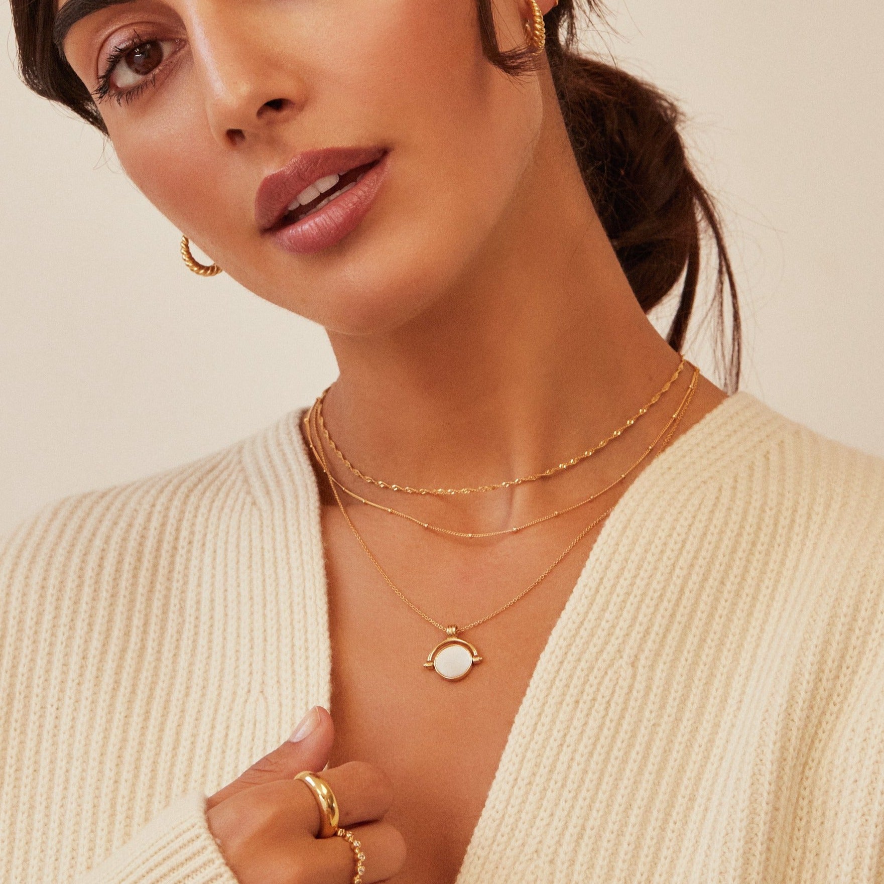 Gold satellite chain necklace layered with a gold mother of pearl spinning disc necklace and a gold twisted rope necklace on a woman wearing a cream knitted jumper holding it open with her hand 