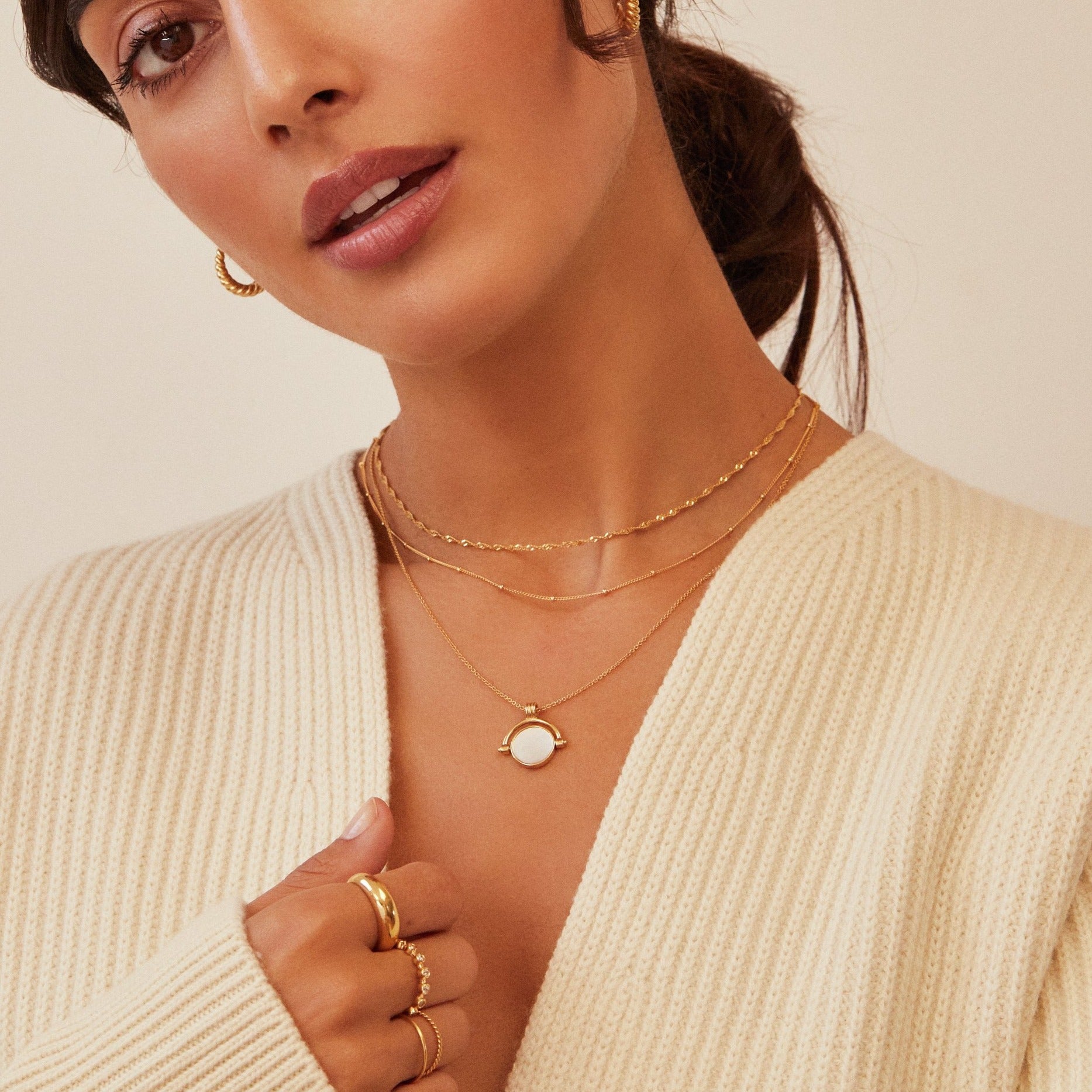 Brunette woman wearing a gold mother of pearl spinning disc necklace layered with a gold satellite chain necklace and a gold twisted rope chain necklace, gold plaine dome ring and other gold rings on her fingers and a cream knitted jumper