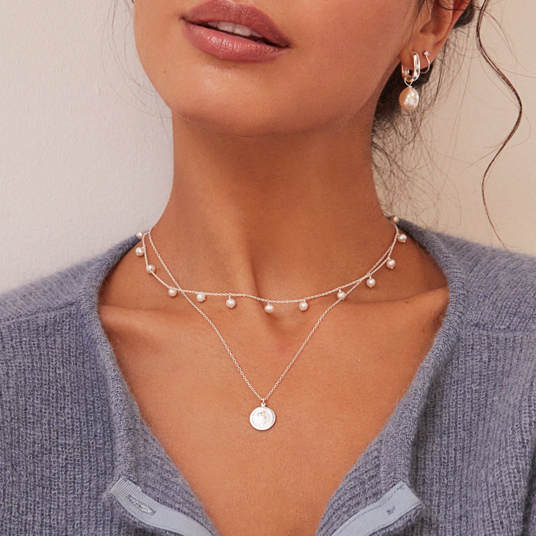 Silver small round St Christopher medallion necklace layered with a silver pearl drop choker around the neck of a woman wearing a silver square edge pearl drop hoop earring in her ear lobe
